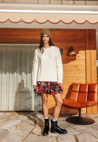 Pull blanc en maille torsadée, jupe fleurie rouge, bottines noires avec chaussettes grises. En arrière-plan, un mur en bois et un mobilier moderne.