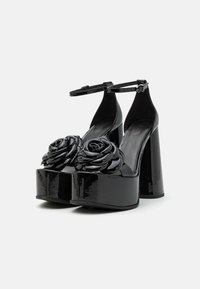 Talons plateformes en cuir verni noir avec de grandes embellissements en forme de rose à l'avant ; elles disposent d'une sangle à la cheville et d'un talon massif.