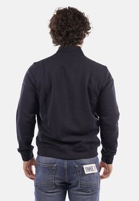 Felpa blu navy con colletto alto, polsini e orlo a costine, realizzata in tessuto liscio. Un dettaglio notevole è l'etichetta a contrasto sulla tasca posteriore.