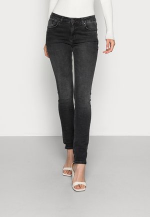 Slim fit zwarte jeans gedragen met een witte geribbelde trui en witte open-teen sandalen met hak tegen een effen achtergrond.