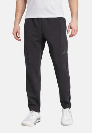 Mann trägt schwarze Sporthose und weiße Turnschuhe, steht mit entspannten Händen an den Seiten vor weißem Hintergrund.