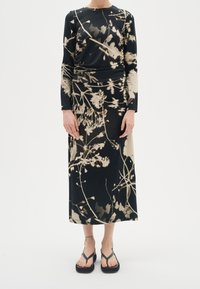 Mujer vistiendo un vestido de manga larga con un estampado floral en negro y beige, con sandalias de plataforma negras, de pie contra un fondo blanco.