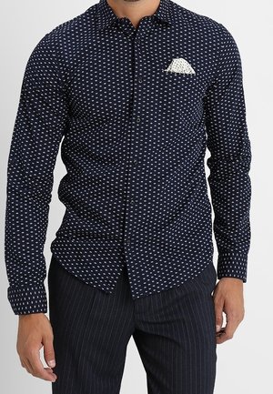 Man draagt een marineblauw overhemd met patroon en witte pochet, gecombineerd met donkere broek met krijtstrepen, staand tegen een witte achtergrond.