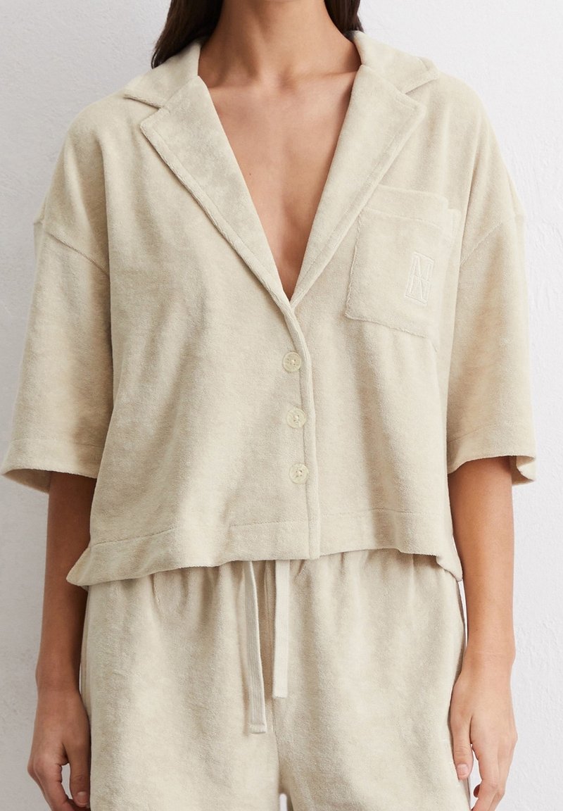 Chemise courte beige, en tissu doux, boutonnée, avec col, manches courtes et poche poitrine ornée d'un détail brodé.