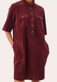 Bordeauxrotes Cordkleid aus Cord mit einem Kragen, durchgehender Knopfleiste, zwei Brusttaschen und Seitentaschen sowie einem halb-lässigen Schnitt.