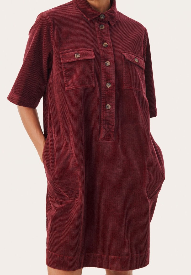 Bordeauxrotes Cordkleid aus Cord mit einem Kragen, durchgehender Knopfleiste, zwei Brusttaschen und Seitentaschen sowie einem halb-lässigen Schnitt.