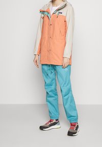 Person wearing a color-blocked hooded jacket in beige and peach, turquoise pants, and gray sneakers with red laces standing on a plain background.