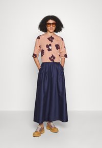 Top cropped com padrão floral em pêssego, combinado com uma saia maxi de cetim azul-marinho. Apresenta decote redondo e mangas largas. Usado com sapatos plataforma em tom de areia.