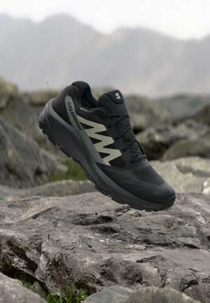 Zapato de senderismo Salomon negro y gris flotando sobre un terreno rocoso con un fondo montañoso borroso.