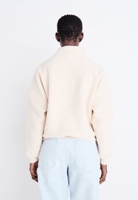 Fleece-Pullover in Creme mit hohem Kragen, lässiger Passform und gerippten Bündchen. Modell kombiniert mit hellblauen Jeans, von hinten betrachtet.