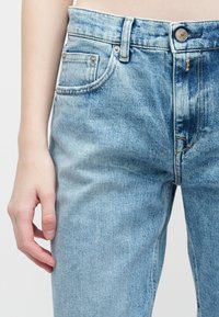 Prim-plan al unei mâini de persoană odihnindu-se lângă corp, îmbrăcată cu blugi de denim de culoare albastru deschis, cu butoane și buzunare vizibile.