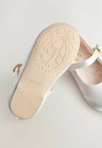 Balerini albe cu o textură netedă și accesorii din aur. Talpa are un design embosat de papuc de balet și dispune de un grip texturat.