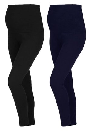 Person, die dunkelblaue Leggings und weiße Turnschuhe trägt, steht mit einem leicht gebeugten Bein vor einem einfarbigen hellen Hintergrund.