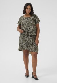 Kurzärmliges Kleid in einem olivgrünen und schwarzen Zebramuster, mit einem lässigen Schnitt und einem dezenten Taillengürtel, getragen mit schwarzen Blockabsatzschuhen.