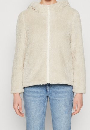 Flødefarvet, blød hættejakke med lynlås, båret med lyseblå denimjeans, vist fra skuldre til hofter.