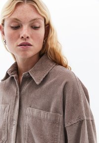 Bruine corduroy shirt met een spitse kraag, knoopsluiting en twee voorzakken; de textuur is ribbelig en zacht, met een relaxte pasvorm.