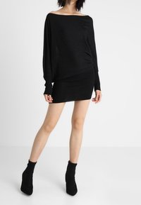 Svart av-axel tröja i kombination med en svart figurnära mini kjol. Tyget verkar mjukt med en lätt ribbad textur. Svarta ankelstövlar fulländar looken.