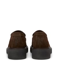 Bruine suède schoenen met een platte zwarte zool, voorzien van een gladde textuur, ronde vorm en subtiele logo-detail aan de achterkant.