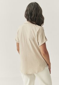 Světle béžová košile s krátkým rukávem a límečkem polo, volný střih a ohrnuté manžety. Měkká textura se rovnými lemy, stylizováno s bílými kalhotami.