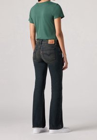 Jean évasé en denim foncé avec poches arrière et écusson logo Levi's ; porté avec un t-shirt vert uni et des chaussures blanches.