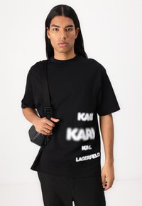 Μαύρο oversize t-shirt από βαμβάκι με διάφορες λευκές επιγραφές "KARL" και πουά μοτίβα. Συνδυασμένο με μια μικρή μαύρη τσάντα ώμου.