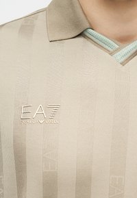 Μπεζ πόλο πουκάμισο με διακριτικά κατακόρυφα λογότυπα EA7 και πράσινη επένδυση στο εσωτερικό του V-λαιμού, φορεμένο από ένα άτομο.