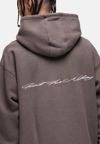 Felpa grigia con cappuccio e vestibilità comoda. Presenta un ricamo bianco in corsivo con la scritta "Good For Nothing" lungo la parte posteriore. Tessuto morbido.