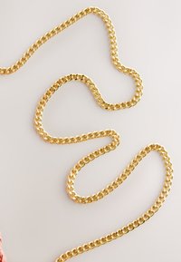 Thick gold chain with interlocking flat links laid out in a loose, curved pattern on a light surface.