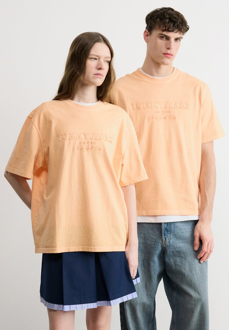 Δύο νέα άτομα φορώντας ίδια ροδακινί μπλουζάκια "Tommy Jeans", στέκονται δίπλα δίπλα μπροστά σε απλό φόντο.