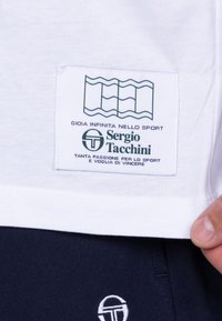 Witte katoenen T-shirt met een groen logo-patch met golvende lijnen, tekst en branding. Korte mouwen en een rechte zoom.