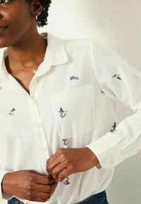 White button-up shirt featuring colorful embroidered figures, including surfers and skiers, with a chest pocket and classic collar.