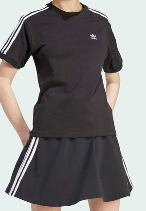 Person trägt ein schwarzes Adidas-T-Shirt mit kurzen Ärmeln und einen passenden Rock mit weißen Streifen an Ärmeln und Rockseiten, steht mit den Händen in den Taschen.