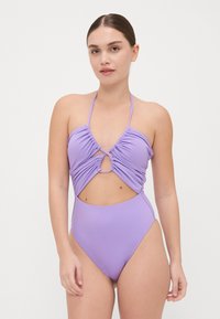 Le maillot de bain une pièce violet présente un bustier froncé, une découpe en forme de trou de serrure au niveau du ventre, des bretelles fines et un design à cuisse haute.