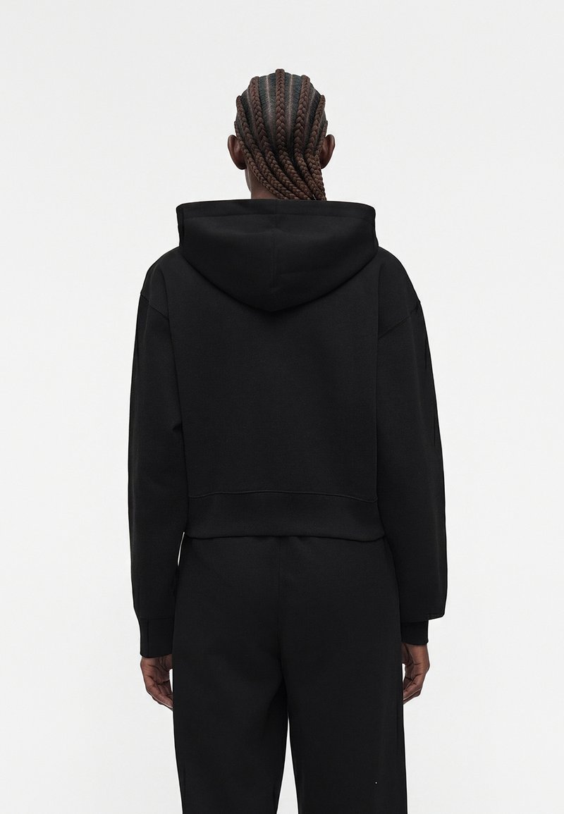 Personne aux cheveux tressés portant un sweat à capuche noir et un pantalon noir, debout de dos sur un fond blanc uni.