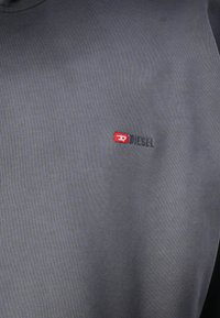 Cămașă gri cu mâneci lungi, cu o textură canalată, având un mic logo roșu "DIESEL" și o patch cu un "D" stilizat pe pieptul stâng.