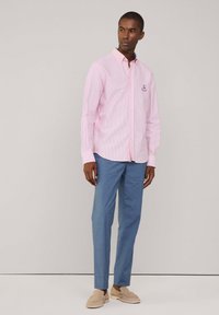 Chemise boutonnée rayée rose et blanche à manches longues, associée à un pantalon bleu clair et des mocassins beiges.