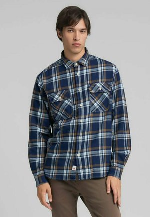 Camisa de franela a cuadros en azul y marrón con collar abotonado, dos bolsillos en el pecho y una pequeña etiqueta con logo en el dobladillo. Tela suave y texturizada.