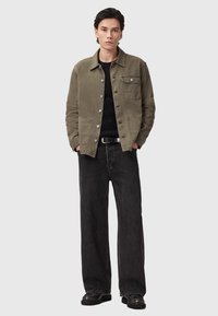 Giacca in canvas verde oliva con bottoni a pressione e tasca sul petto, abbinata a jeans neri a gamba larga e stivali neri; maglione a maglia strutturata sotto.