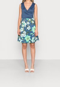 Blå ärmlös klänning med en omslagdesign, med ett blommönster i grönt och vitt; knä-lång kjol; bärs med gräddfärgade öppna skor.