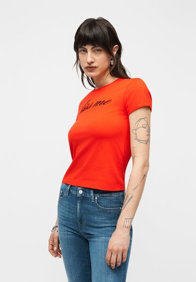 Γυναίκα με σκούρα μαλλιά που φοράει φωτεινό πορτοκαλί κοντομάνικο τοπ με το κείμενο «Μου λείπεις» και μπλε τζιν, δείχνοντας τα τατουάζ στο αριστερό της χέρι.