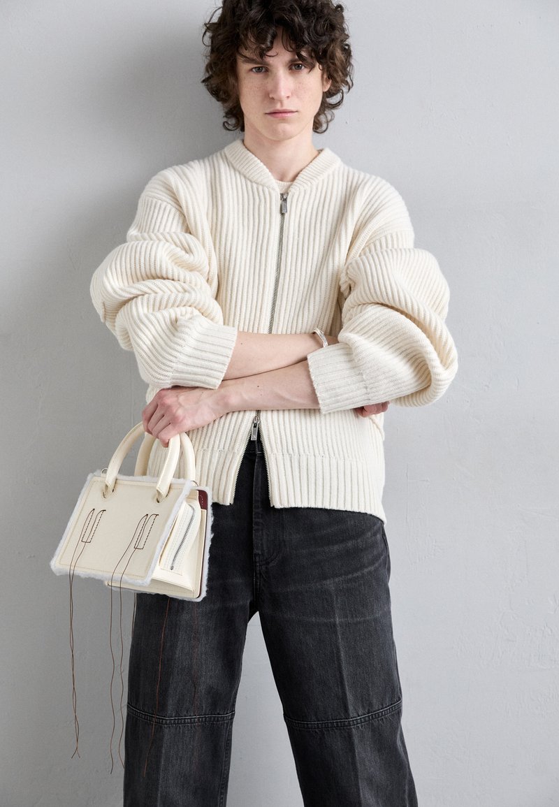 Krémová pruhovaná pletená cardigan s predným zipsom, v kombinácii s tmavými džínsami a malou krémovou kabelkou s jedinečným šitím a akcentmi.