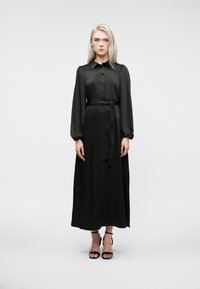 Naine seisab valge tausta ees, seljas pikk must satiinist kleit, millel on puffed pikkade varrukatega ning jalas mustad pahkluu rihmadega kingad.