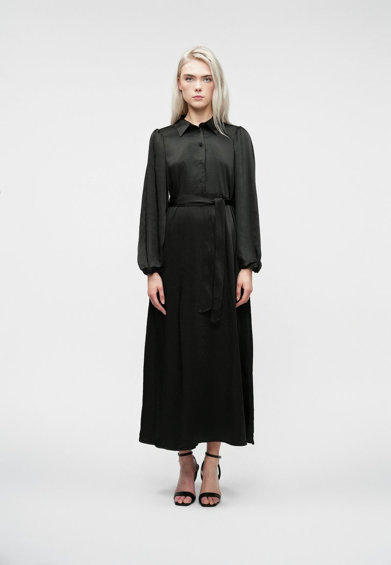 Naine seisab valge tausta ees, seljas pikk must satiinist kleit, millel on puffed pikkade varrukatega ning jalas mustad pahkluu rihmadega kingad.