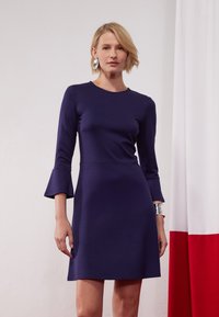 Rochie lungă bleumarin cu mâneci clopot, realizată dintr-un material neted, având un guler rotund clasic. Accent cu brățară argintie pe încheietura mâinii.