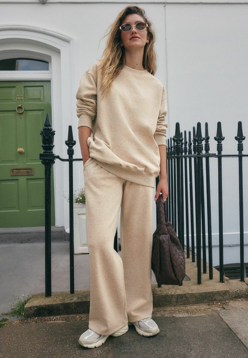 Beige, strukturiertes Sweatshirt und passende weite Hosen. Getragen mit grauen Sneakers und einer braunen geflochtenen Tasche. Übergroße Sonnenbrillen runden den Look ab.
