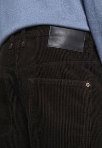 Pantaloni in corduroy marrone scuro con righe verticali, caratterizzati da un'etichetta in pelle nera e dettagli sulle tasche frontali. Il tessuto testurizzato aggiunge profondità.