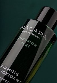 Svart cylindrisk flaska med glansig yta. Etiketten visar "MÁDARA" och "Infusion Vert" i silver. Produkten är en fastgörande kroppsolja.