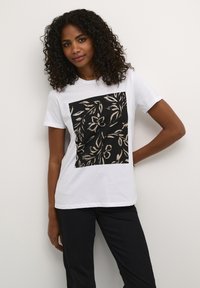 T-shirt en coton blanc avec un motif graphique carré noir orné de fleurs beiges. Manches courtes, encolure ras du cou, coupe décontractée.