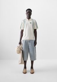 Beige short-sleeve polo with green stripes, denim shorts, and beige slip-on shoes. Holding a beige bag and a canvas tote.