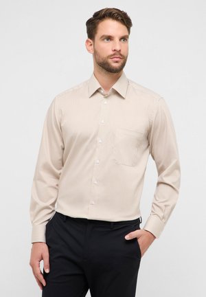 Beigefarbenes gestreiftes Hemd mit langen Ärmeln, geknöpftem Kragen und einer einzelnen Brusttasche. Der Stoff hat eine glatte Textur. Kombiniert mit dunkler Hose.